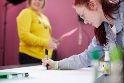 Female student with red hair and clear glasses using a Sharpie to write on a notepad with classmate in background
