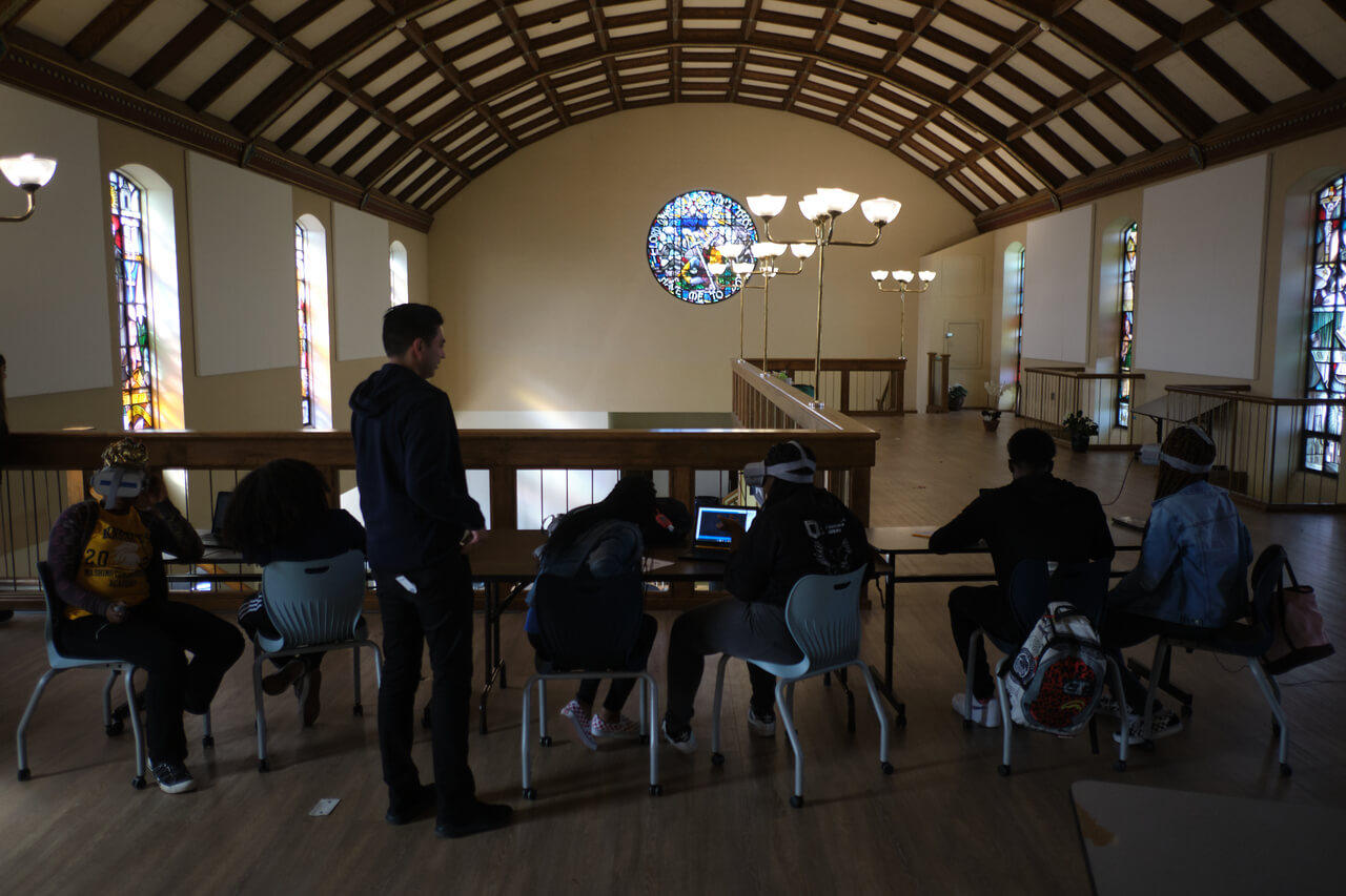 Landscape view of seven students sitting in a large room with beamed ceilings and stained glass windows practicing VR