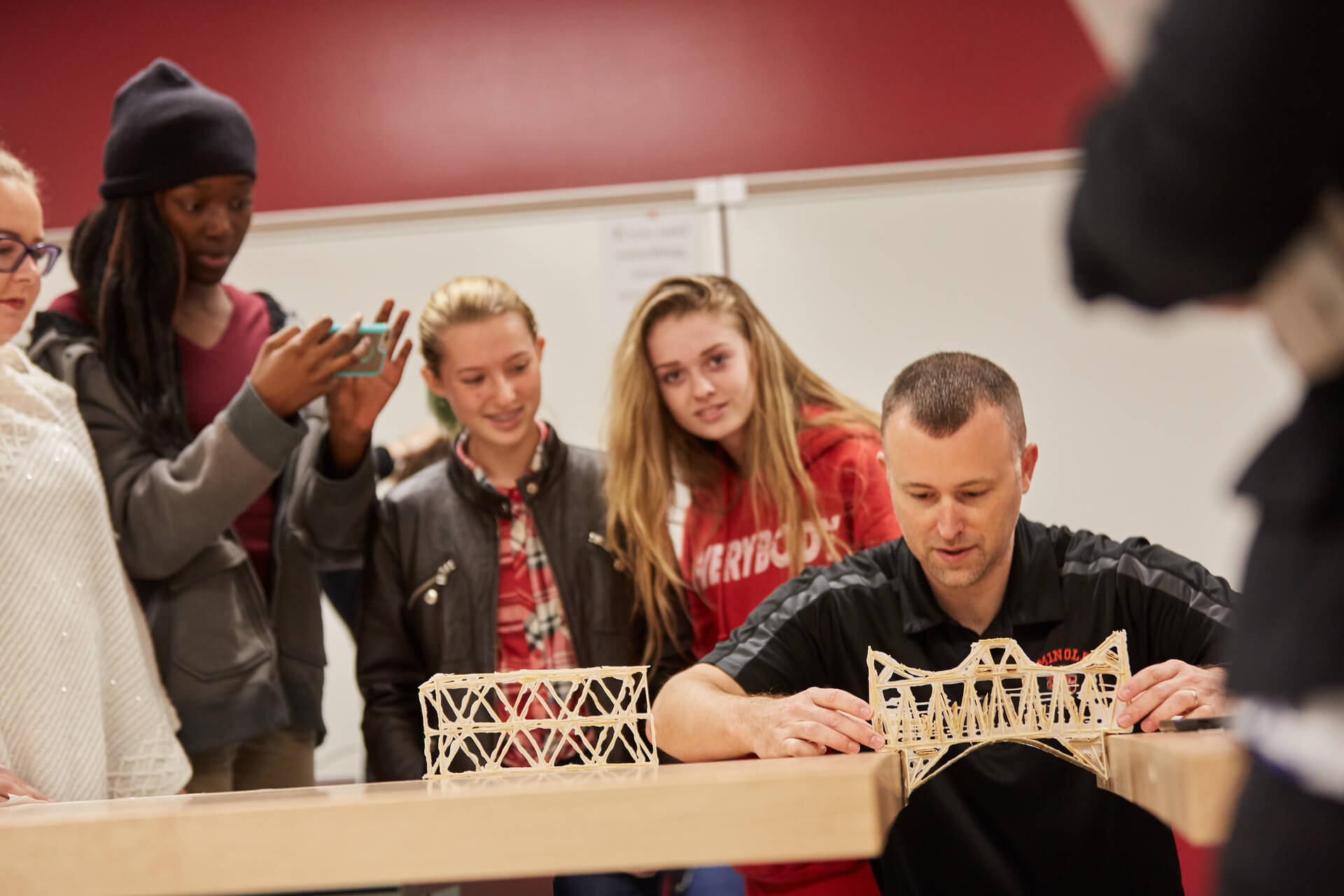 Teacher testing the strength of a student-made toothpick bridge with students watching in the background