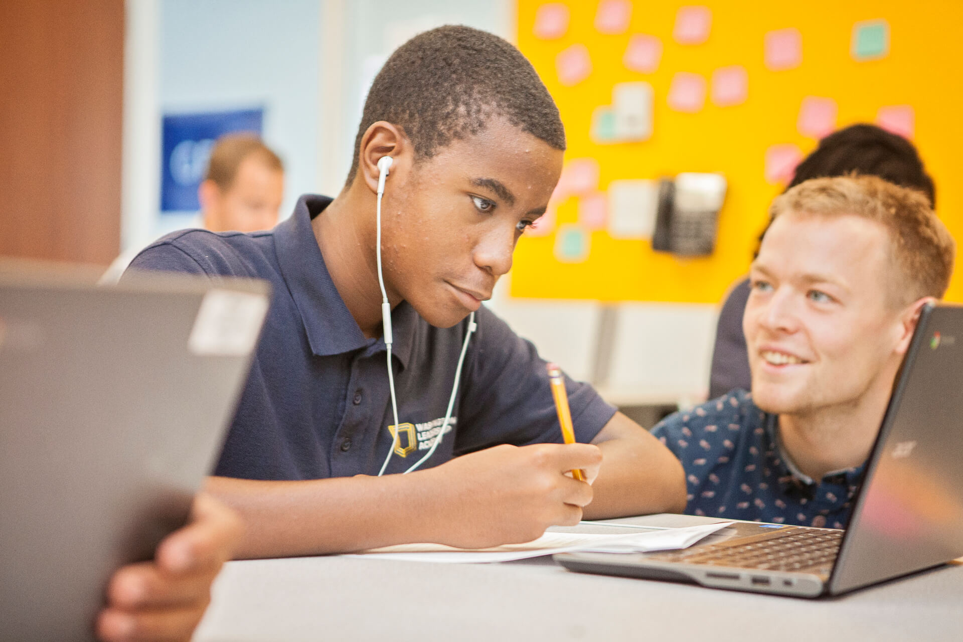Student wearing headphones and taking notes in front of their computer with another student smiling in the background