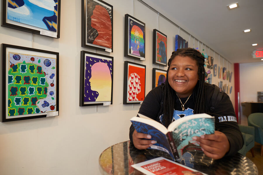 A student in a black hoodie reads a graphic novel and smiles at student artwork displayed in the classroom.